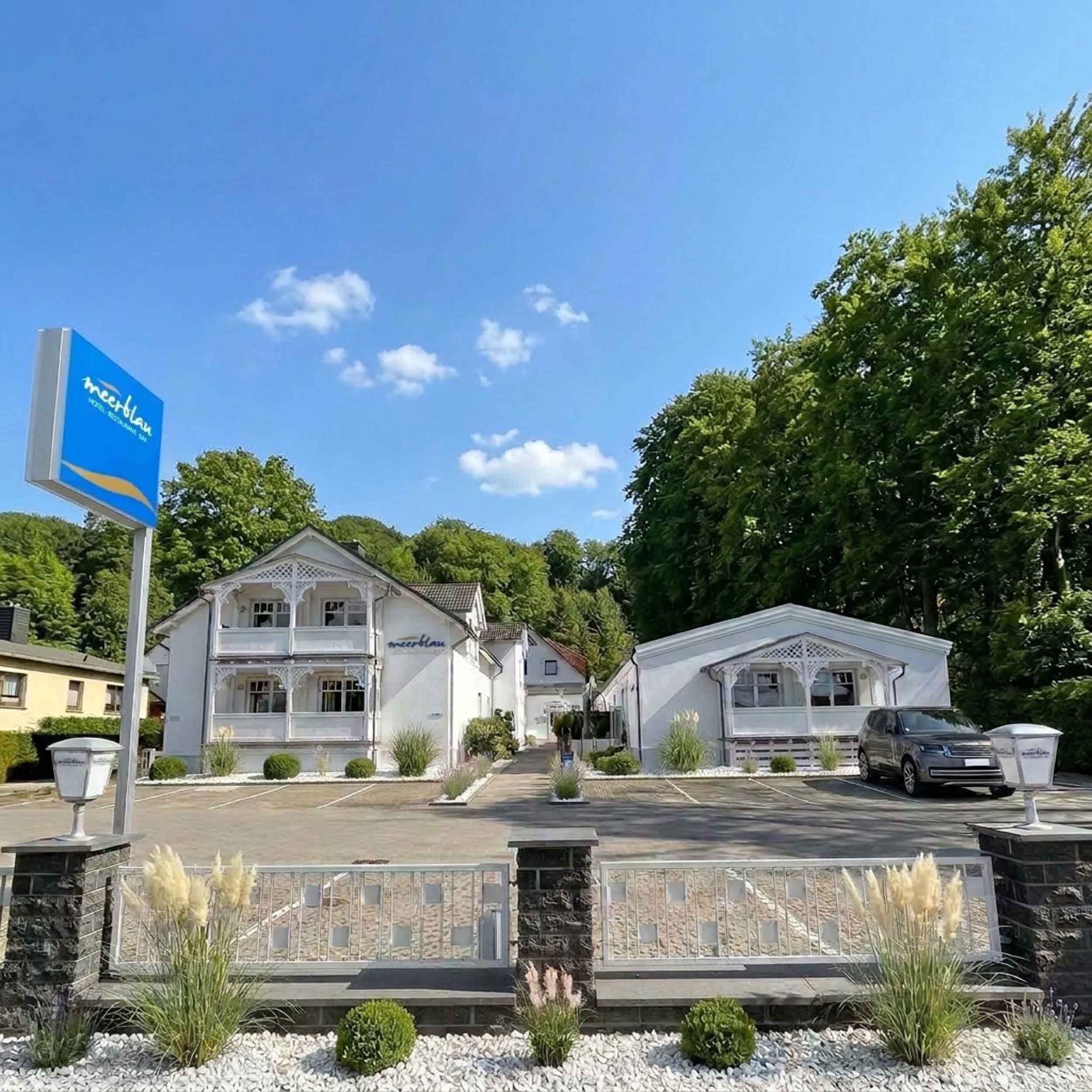 Hotel meerblau im Ostseebad Sellin auf Rügen: weiße Häuser in Bäderarchitektur mit verzierten Holzbalkonen, Hotelschild und Parkplatz vor Buchenwald